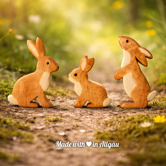 Handgefertigtes Holzspielzeug Feldhasen-Familie von Allgäuer Holztiere mit Häsin, Rammler und Hasenbaby, auf einem natürlichen Wald- und Wiesenweg arrangiert, umgeben von Moos und kleinen Blüten, vor einer unscharfen sonnigen Naturlandschaft, aus bayerischem Ahornholz gefertigt, mit pflanzlichen Farben in Braun-, Hellbraun- und Naturtönen bemalt, im Allgäu produziert und für Kinder ab 3 Jahren geeignet.