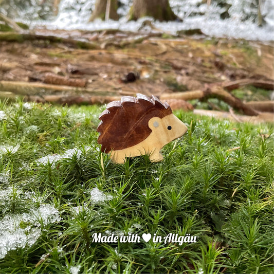 Handgefertigtes Holzspielzeug Igel Baby stehend von Allgäuer Holztiere, auf moosigem Waldboden mit etwas Schnee, im Hintergrund unscharfe Baumstämme, aus bayerischem Ahornholz gefertigt, braun und grau mit pflanzlichen Farben bemalt, nachhaltig im Allgäu produziert und für Kinder ab 3 Jahren geeignet.