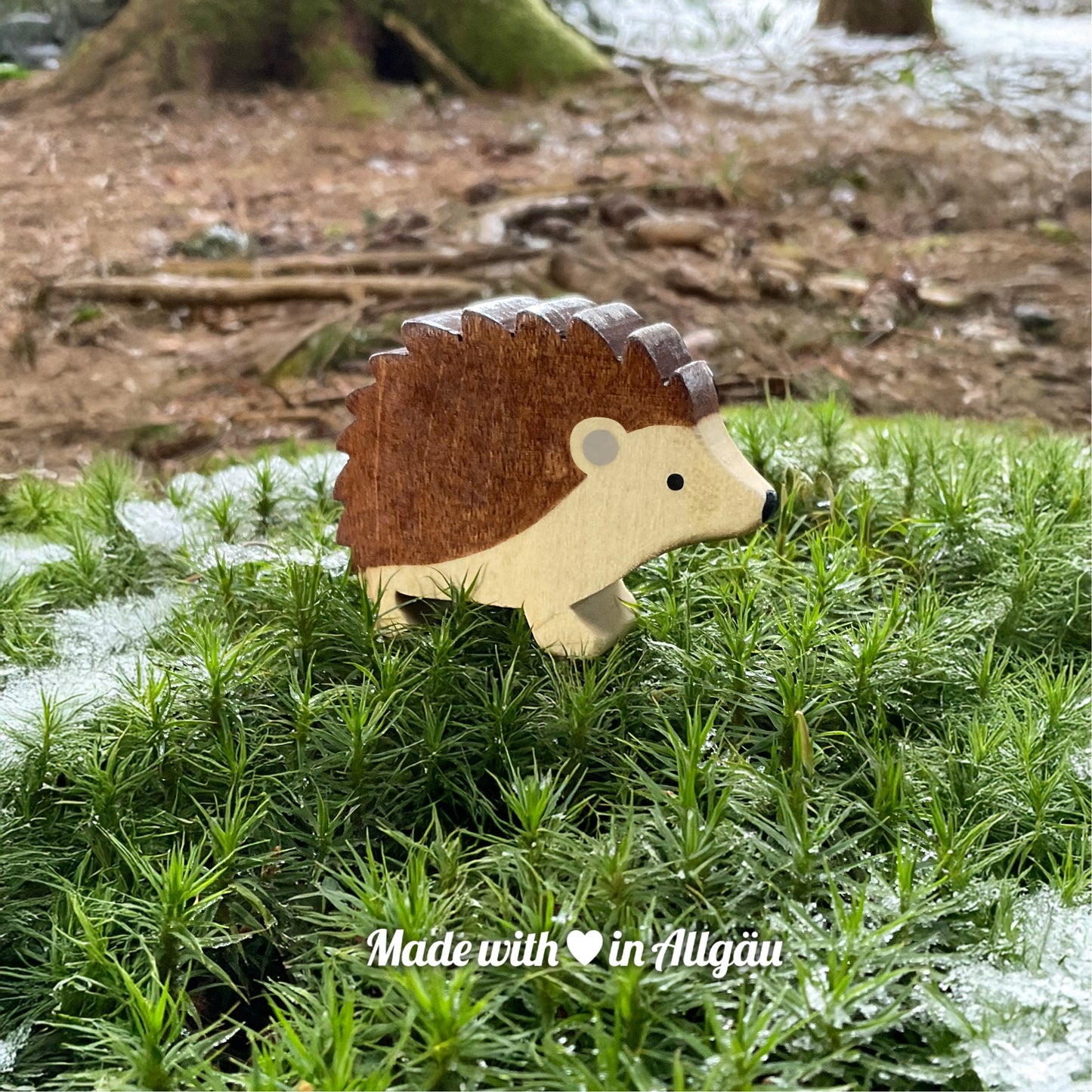 Handgefertigtes Holzspielzeug Igel Männchen stehend von Allgäuer Holztiere, auf moosigem Waldboden mit etwas Schnee, im Hintergrund unscharfe Äste und Tannenzapfen, aus bayerischem Ahornholz gefertigt, braun und grau mit pflanzlichen Farben bemalt, nachhaltig im Allgäu produziert und für Kinder ab 3 Jahren geeignet.