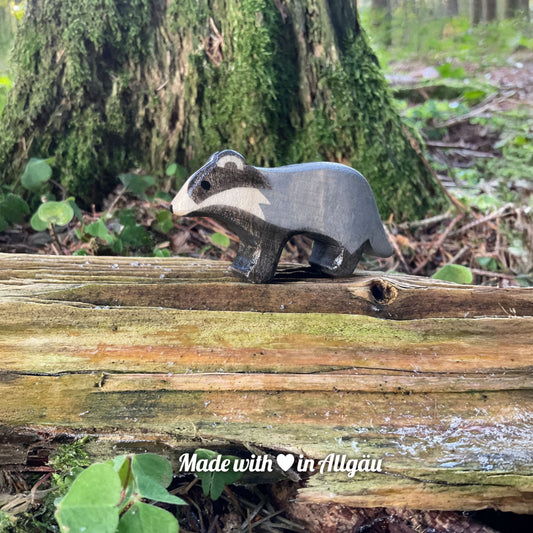 Handgefertigtes Holzspielzeug Dachs-Baby grau stehend von Allgäuer Holztiere, auf bemoostem, verwittertem Holz in natürlicher Waldlandschaft platziert, aus bayerischem Ahornholz gefertigt, grau und schwarz mit pflanzlichen Farben bemalt, nachhaltig im Allgäu produziert und für Kinder ab 3 Jahren geeignet.