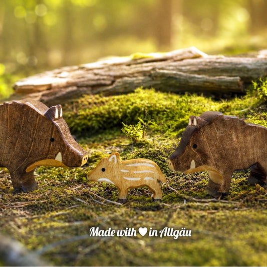 Handgefertigtes Holzspielzeug Wildschwein-Familie von Allgäuer Holztiere mit Keiler, Bache und Frischling, auf einem moosbedeckten Waldboden vor einem Baumstamm arrangiert, umgeben von natürlichen Waldmaterialien, im Hintergrund eine unscharfe Waldlandschaft, aus bayerischem Ahornholz gefertigt, mit pflanzlichen Farben in Braun-, Hellbraun- und Schwarztönen bemalt, nachhaltig im Allgäu produziert und für Kinder ab 3 Jahren geeignet.
