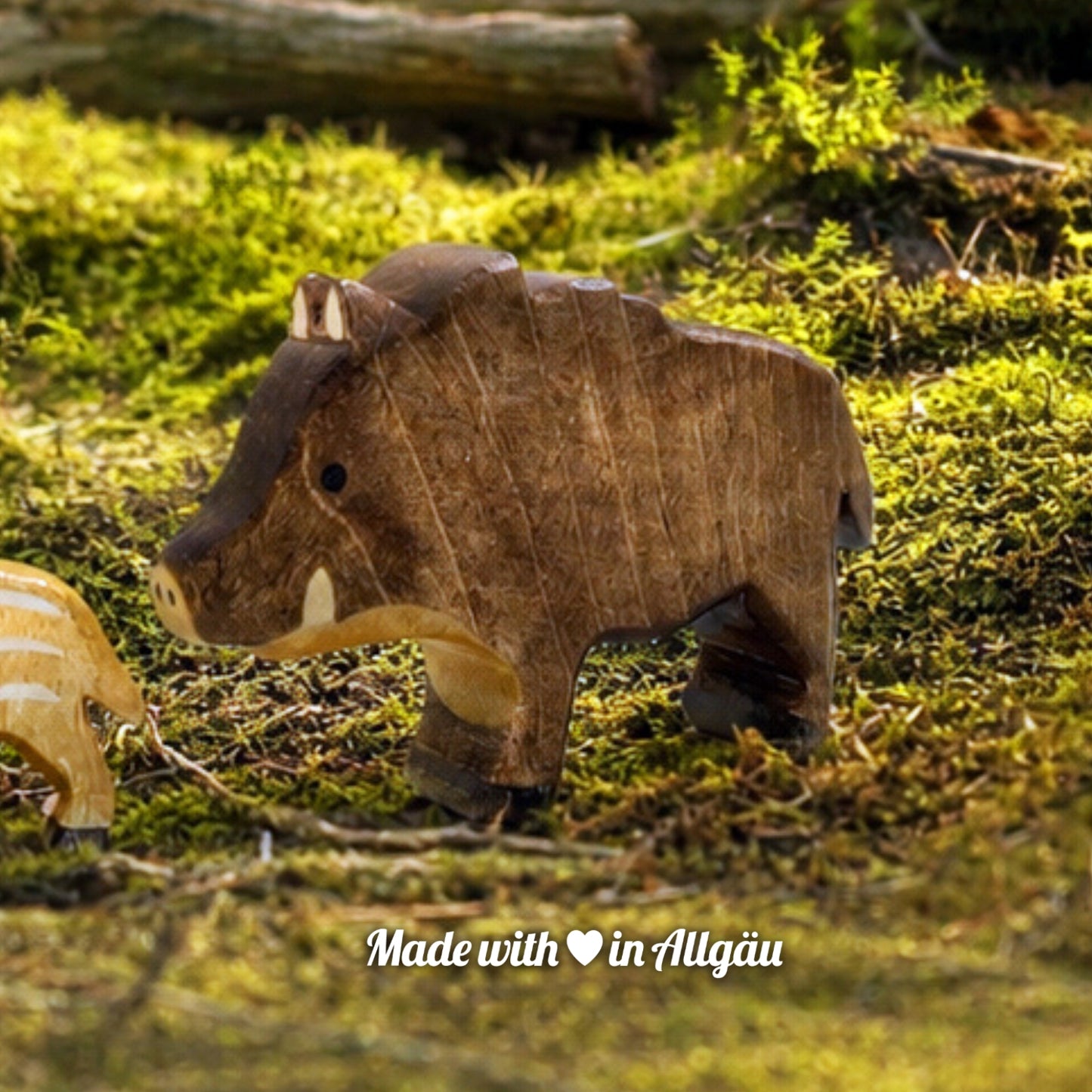 Handgefertigtes Holzspielzeug Wildschwein Bache von Allgäuer Holztiere, auf einem moosbedeckten Waldboden vor einem Baumstamm stehend, umgeben von natürlichen Waldmaterialien, im Hintergrund eine unscharfe Waldlandschaft, aus bayerischem Ahornholz gefertigt, mit pflanzlichen Farben braun, hellbraun und schwarz bemalt, nachhaltig im Allgäu produziert und für Kinder ab 3 Jahren geeignet.