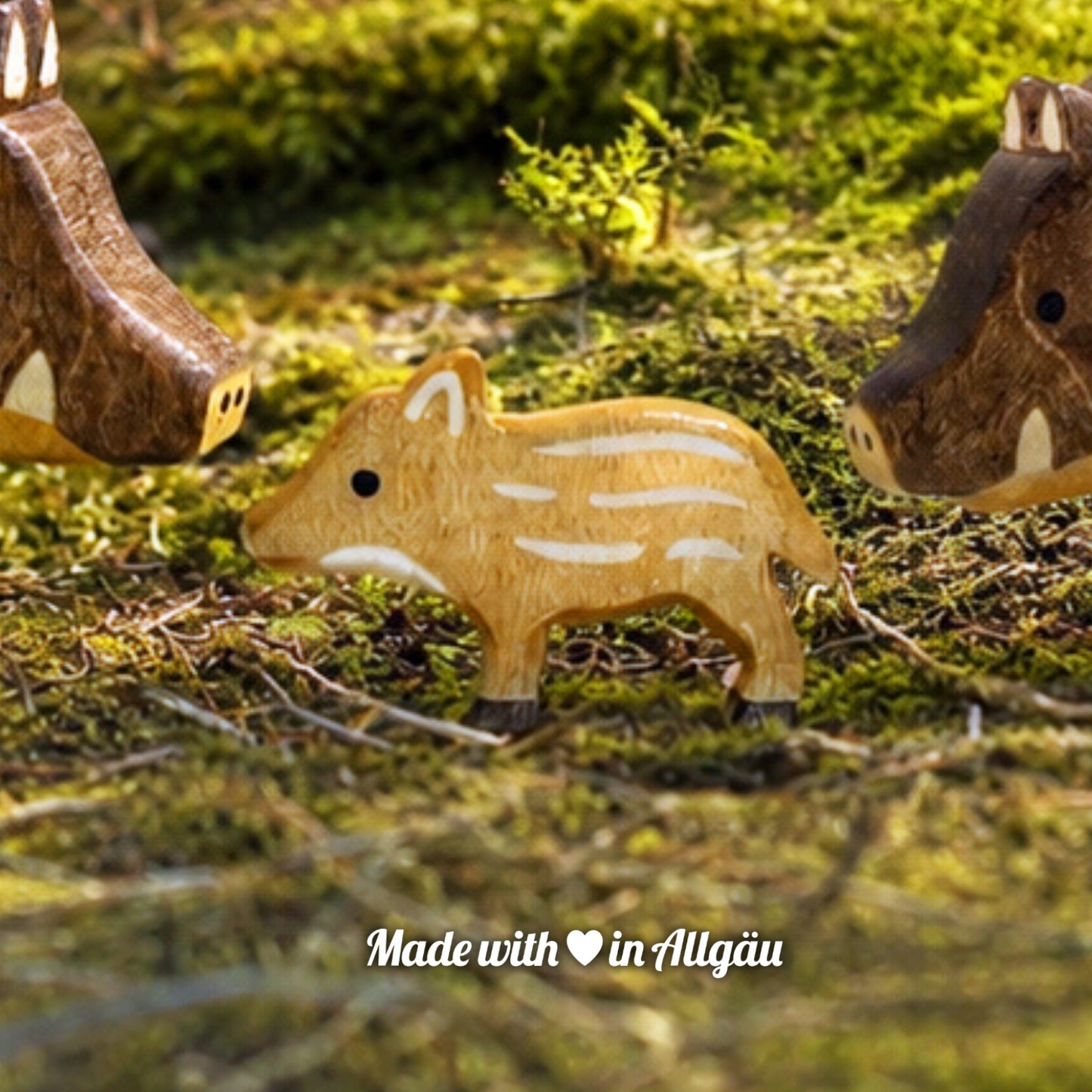 Handgefertigtes Holzspielzeug Wildschwein Frischling von Allgäuer Holztiere, auf einem moosbedeckten Waldboden zwischen einem Wildschwein-Keiler und einer Wildschwein-Bache stehend, umgeben von natürlichen Waldmaterialien, im Hintergrund eine unscharfe Waldlandschaft, aus bayerischem Ahornholz gefertigt, mit pflanzlichen Farben hellbraun und schwarz bemalt, nachhaltig im Allgäu produziert und für Kinder ab 3 Jahren geeignet.