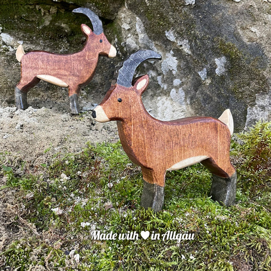 Handgefertigtes Holzspielzeug Steinbock Bock von Allgäuer Holztiere, stehend auf Moos und Felsen in natürlicher Berglandschaft der Alpen, aus bayerischem Ahornholz gefertigt, braun und schwarz bemalt mit pflanzlichen Farben, nachhaltig produziert im Allgäu und für Kinder ab 3 Jahren geeignet.