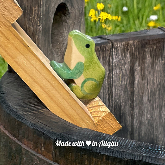 Handgefertigtes Holzspielzeug Frosch Weibchen von Allgäuer Holztiere, hellgrün und dunkelgrün bemalt mit pflanzlichen Farben, sitzend in Regentonne oder natürlicher Uferlandschaft, aus bayerischem Ahornholz gefertigt, nachhaltig produziert im Allgäu und für Kinder ab 3 Jahren geeignet.