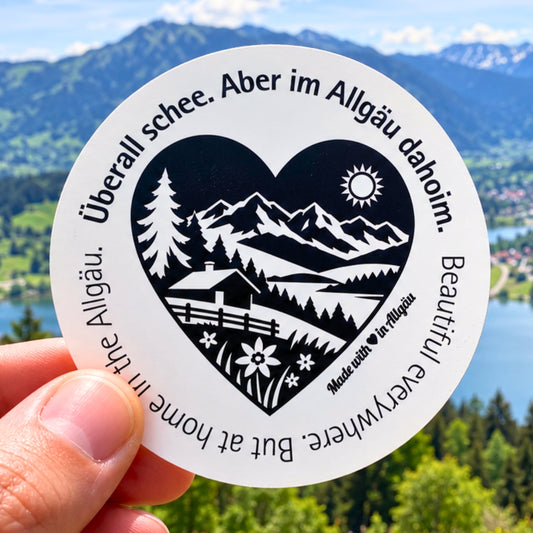 Runder Allgäu-Aufkleber „Überall schee. Aber im Allgäu dahoim.“ mit schwarz-weißem Herzmotiv, Bergpanorama, Tannen, Alpe und Blumenwiese, vor dem Großen Alpsee bei Immenstadt. Ein Heimat-Sticker als Allgäu-Souvenir, Urlaubserinnerung und für liebe Grüße aus der Ferne.