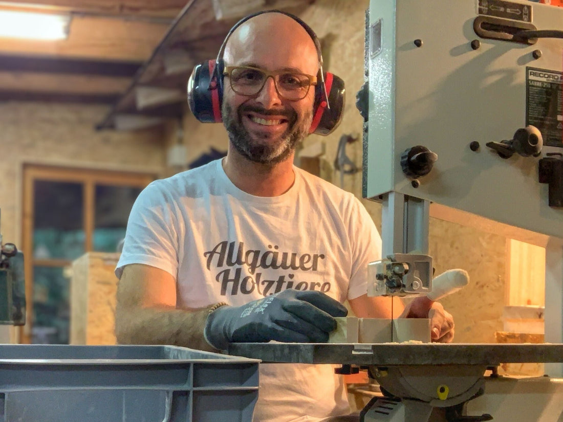 Mann mit Gehörschutz sägt Holz an einer Bandsäge in Werkstatt, trägt T-Shirt mit „Allgäuer Holztier“ und lächelt in die Kamera.