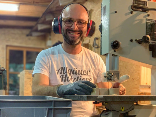 Mann mit Gehörschutz sägt Holz an einer Bandsäge in Werkstatt, trägt T-Shirt mit „Allgäuer Holztier“ und lächelt in die Kamera.