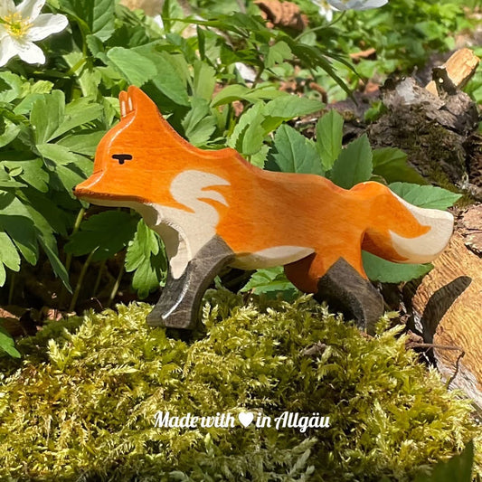 Handgefertigtes Holzspielzeug Fuchs Fähe rot von Allgäuer Holztiere, orange und schwarz bemalt mit pflanzlichen Farben, stehend in natürlicher Umgebung auf Waldboden oder Moos, aus bayerischem Ahornholz gefertigt, nachhaltig produziert im Allgäu und für Kinder ab 3 Jahren geeignet.