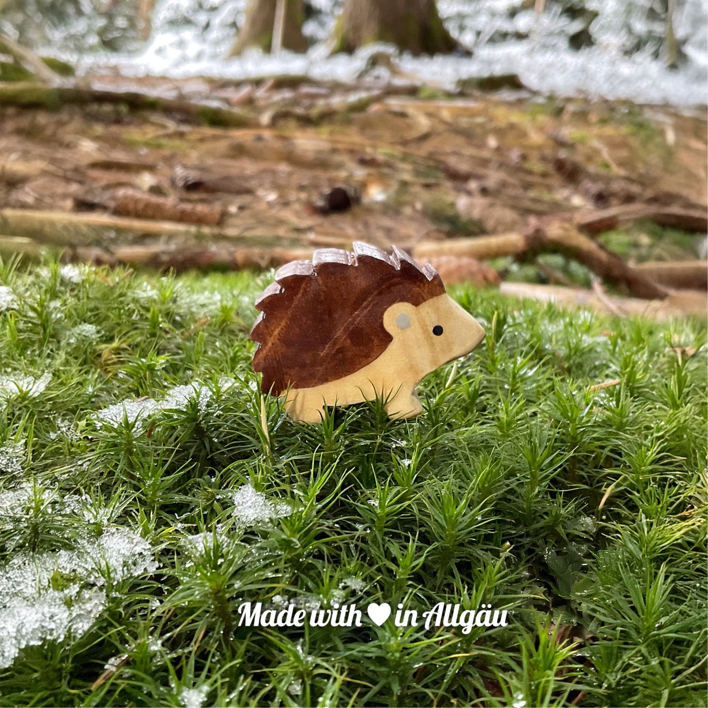 Handgefertigtes Holzspielzeug Igel Baby stehend von Allgäuer Holztiere, auf moosigem Waldboden mit etwas Schnee, im Hintergrund unscharfe Baumstämme, aus bayerischem Ahornholz gefertigt, braun und grau mit pflanzlichen Farben bemalt, nachhaltig im Allgäu produziert und für Kinder ab 3 Jahren geeignet.