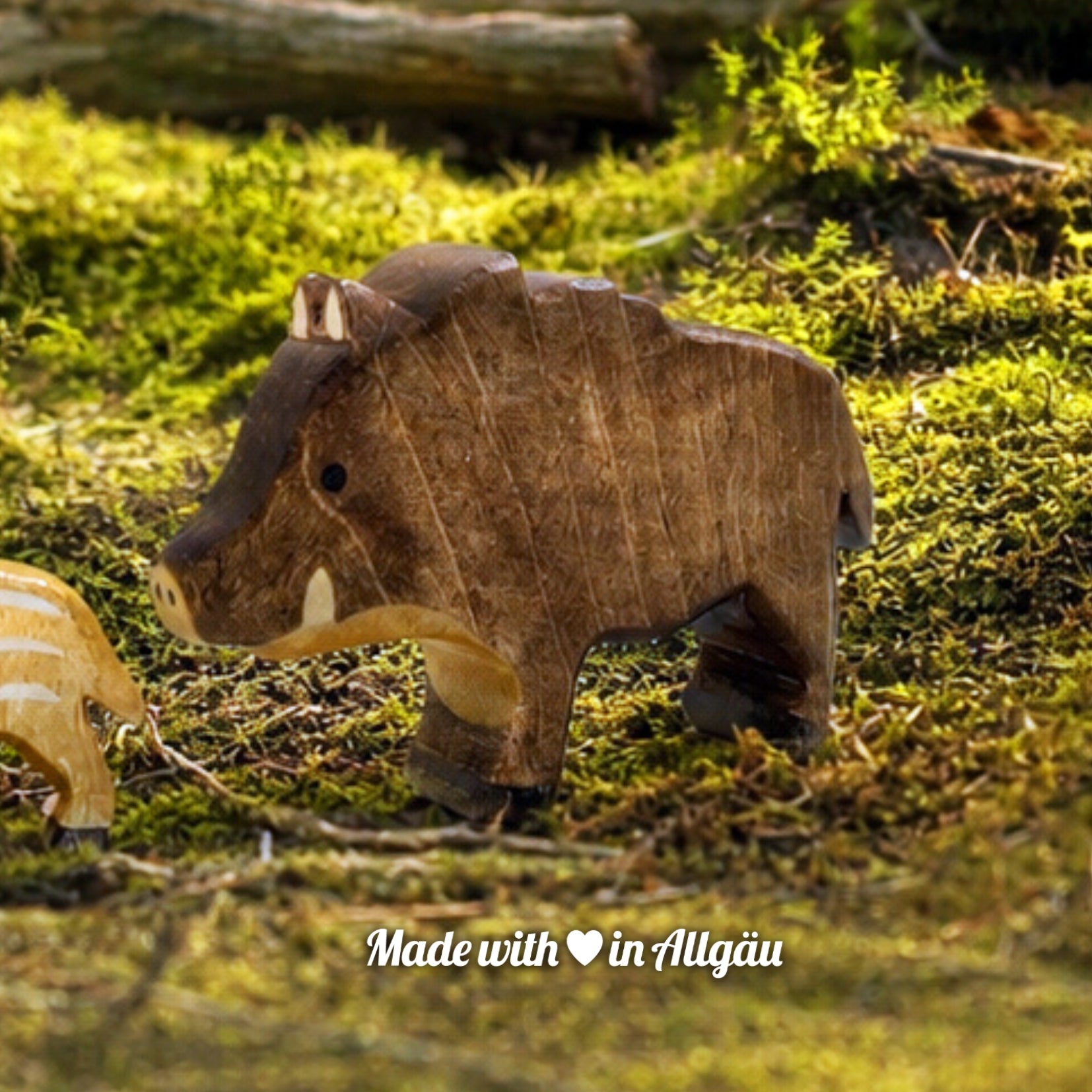 Handgefertigtes Holzspielzeug Wildschwein Bache von Allgäuer Holztiere, auf einem moosbedeckten Waldboden vor einem Baumstamm stehend, umgeben von natürlichen Waldmaterialien, im Hintergrund eine unscharfe Waldlandschaft, aus bayerischem Ahornholz gefertigt, mit pflanzlichen Farben braun, hellbraun und schwarz bemalt, nachhaltig im Allgäu produziert und für Kinder ab 3 Jahren geeignet.