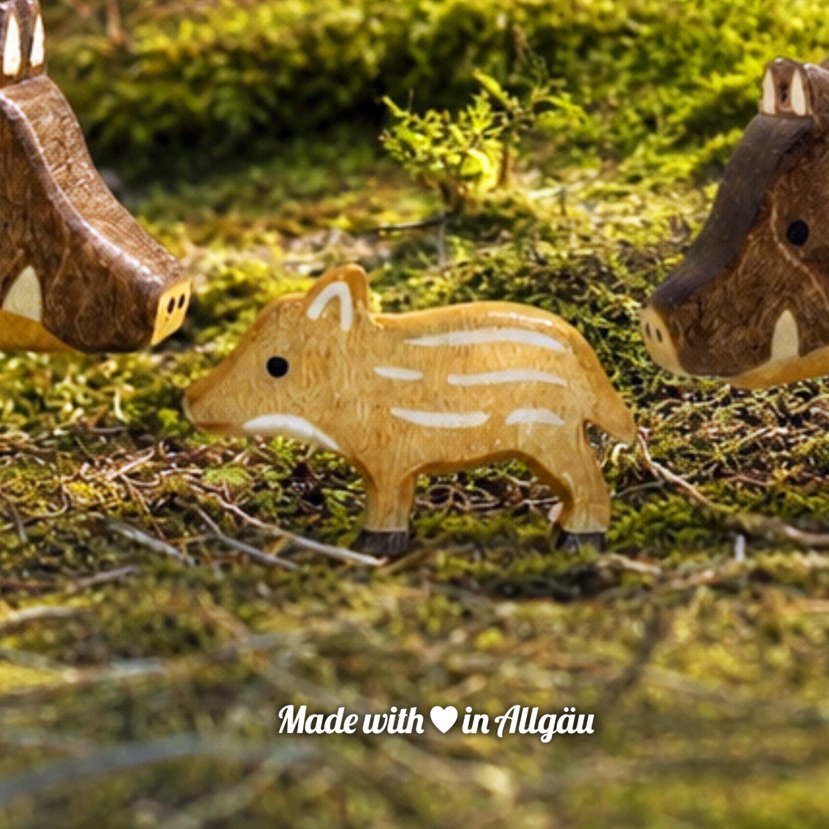 Handgefertigtes Holzspielzeug Wildschwein Frischling von Allgäuer Holztiere, auf einem moosbedeckten Waldboden zwischen einem Wildschwein-Keiler und einer Wildschwein-Bache stehend, umgeben von natürlichen Waldmaterialien, im Hintergrund eine unscharfe Waldlandschaft, aus bayerischem Ahornholz gefertigt, mit pflanzlichen Farben hellbraun und schwarz bemalt, nachhaltig im Allgäu produziert und für Kinder ab 3 Jahren geeignet.