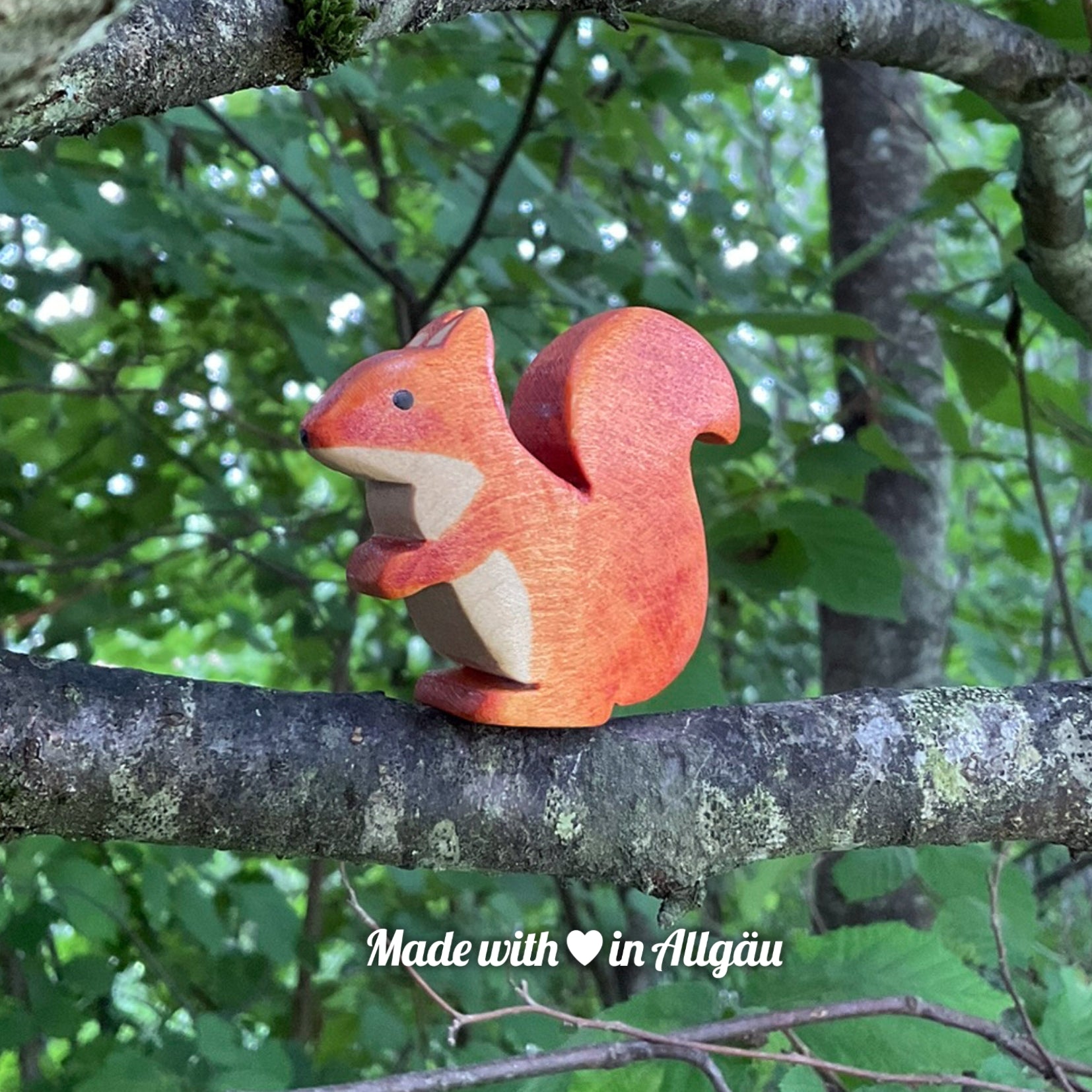 Handgefertigtes Holzspielzeug Eichhörnchen weiblich von Allgäuer Holztiere, braun bemalt mit pflanzlichen Farben, sitzend auf einem Ast in natürlicher Umgebung, aus bayerischem Ahornholz gefertigt, nachhaltig produziert im Allgäu und für Kinder ab 3 Jahren geeignet.