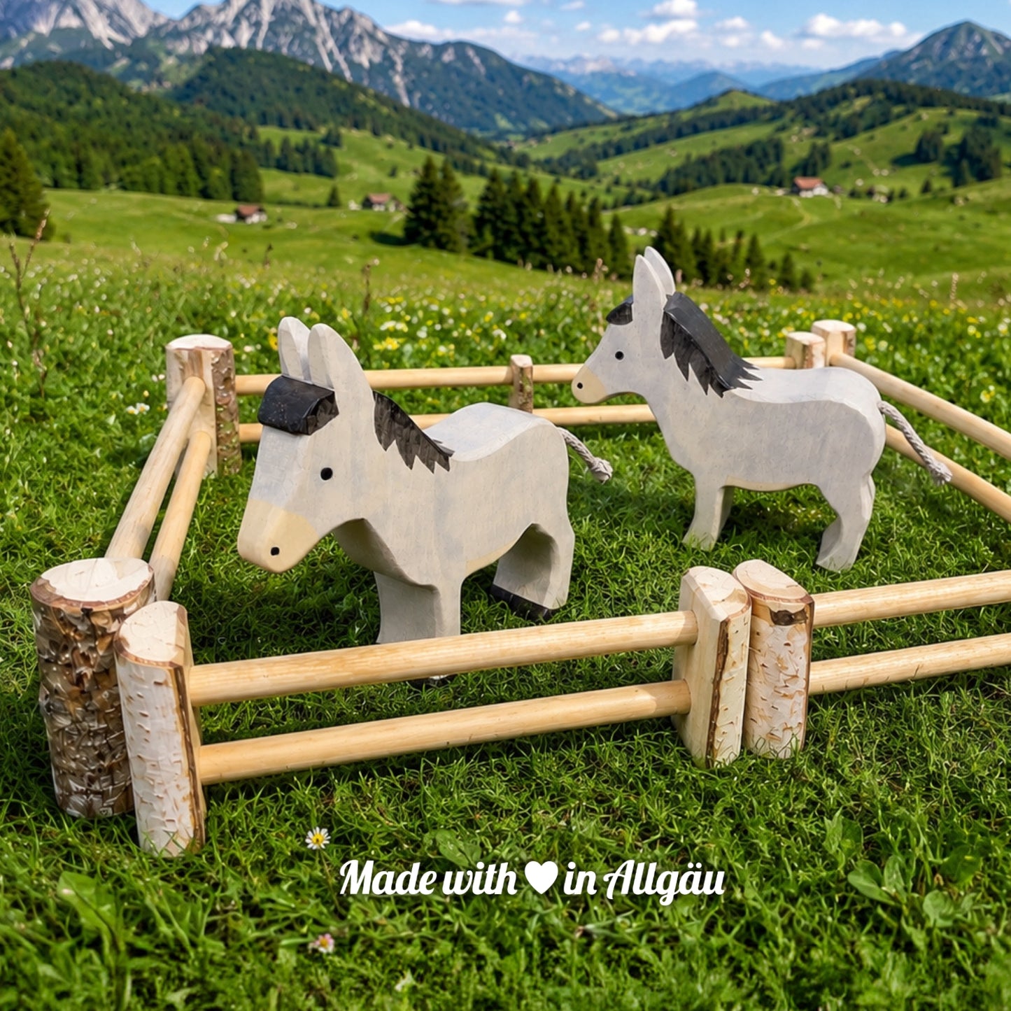 Handgefertigtes Holzspielzeug Esel Stute grau stehend von Allgäuer Holztiere, auf einer grünen Bergwiese mit Holzzaun und Alpenpanorama, aus bayerischem Ahornholz gefertigt, grau und schwarz bemalt mit pflanzlichen Farben, produziert im Allgäu und für Kinder ab 3 Jahren geeignet.