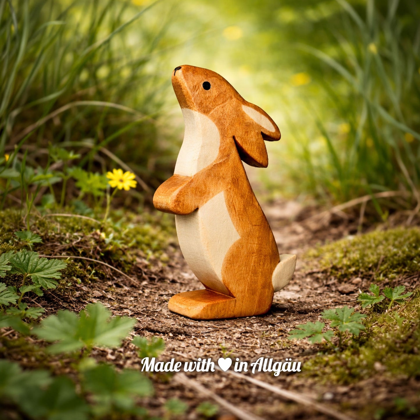 Handgefertigtes Holzspielzeug Feldhase  Rammler  Männchen von Allgäuer Holztiere, auf einem Waldweg zwischen Gras und kleinen Blumen stehend, im Hintergrund unscharfe grüne Vegetation, aus bayerischem Ahornholz gefertigt, mit pflanzlichen Farben braun und schwarz bemalt, nachhaltig im Allgäu produziert und für Kinder ab 3 Jahren geeignet.