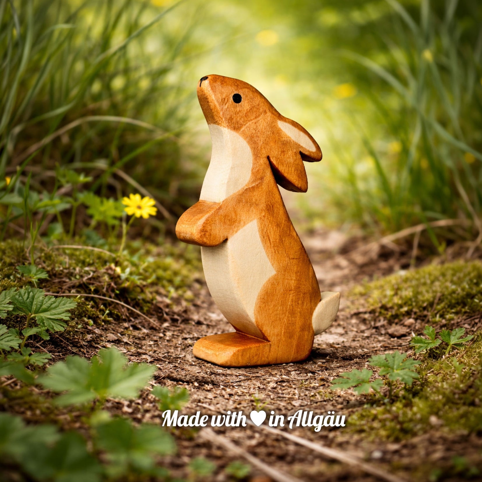 Handgefertigtes Holzspielzeug Feldhase  Rammler  Männchen von Allgäuer Holztiere, auf einem Waldweg zwischen Gras und kleinen Blumen stehend, im Hintergrund unscharfe grüne Vegetation, aus bayerischem Ahornholz gefertigt, mit pflanzlichen Farben braun und schwarz bemalt, nachhaltig im Allgäu produziert und für Kinder ab 3 Jahren geeignet.