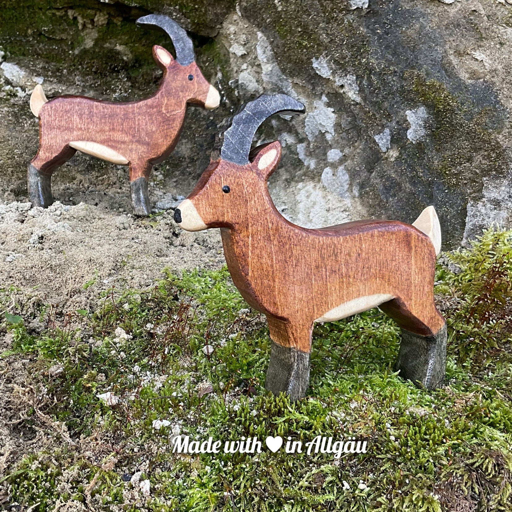 Handgefertigtes Holzspielzeug Steinbock Bock von Allgäuer Holztiere, stehend auf Moos und Felsen in natürlicher Berglandschaft der Alpen, aus bayerischem Ahornholz gefertigt, braun und schwarz bemalt mit pflanzlichen Farben, nachhaltig produziert im Allgäu und für Kinder ab 3 Jahren geeignet.