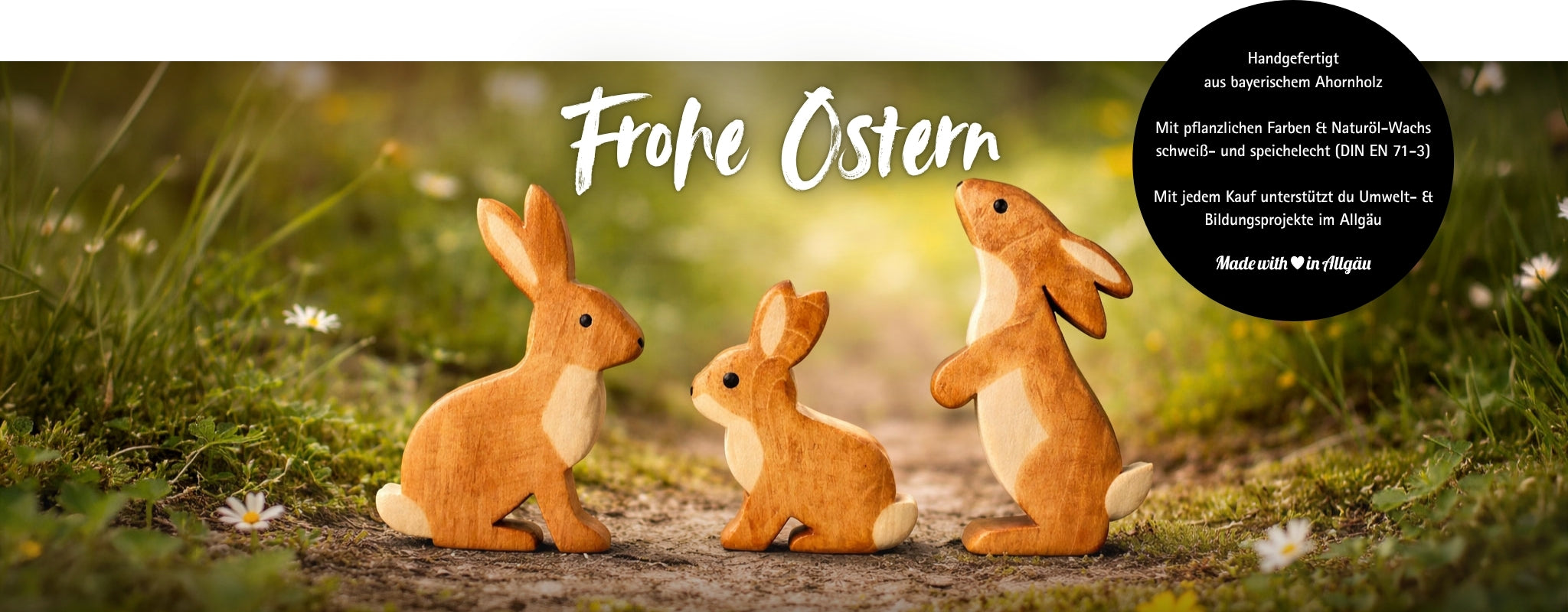 Handgefertigte Holzhasen von Allgäuer Holztiere aus bayerischem Ahornholz für offenes Speil oder als nachhaltige Osterdeko, drei braune Hasenfiguren auf Naturweg mit Blumen und Schriftzug „Frohe Ostern“, bemalt mit pflanzlichen Farben und mit Naturöl-Wachs geschützt, Made with Love in Allgäu