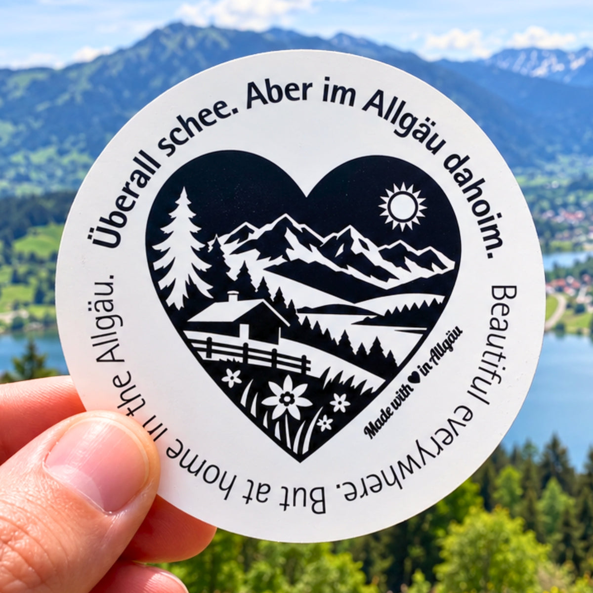 Runder Allgäu-Aufkleber „Überall schee. Aber im Allgäu dahoim.“ mit schwarz-weißem Herzmotiv, Bergpanorama, Tannen, Alpe und Blumenwiese, vor dem Großen Alpsee bei Immenstadt. Ein Heimat-Sticker als Allgäu-Souvenir, Urlaubserinnerung und für liebe Grüße aus der Ferne.
